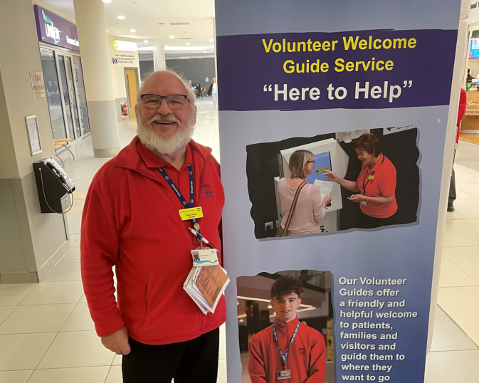 Volunteer David Henry standing next to a volunteering sign in the atrium of the Queen Elizabeth University Hospitail